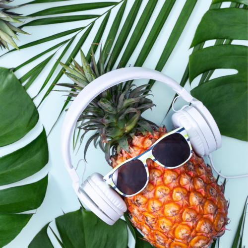 A pineapple wearing white headphones and a pair of white-rimmed sunglasses, is tilted against a background of green tropical leaves on a pale blue surface. surface.